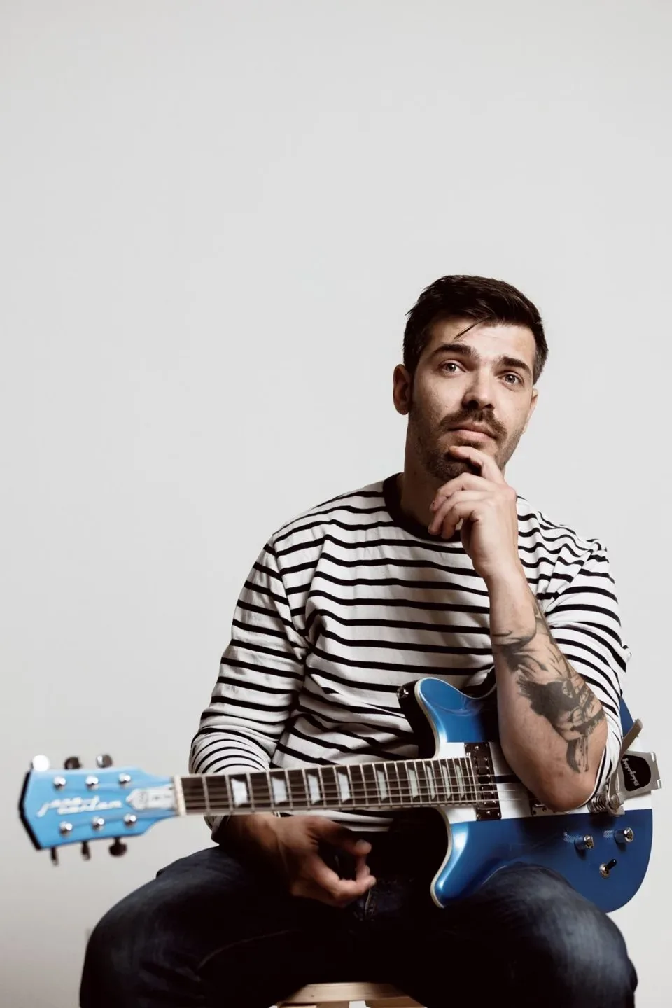 Matteo Leone con la maglia a righe da marinaio e la chitarra blu, mano sul mento