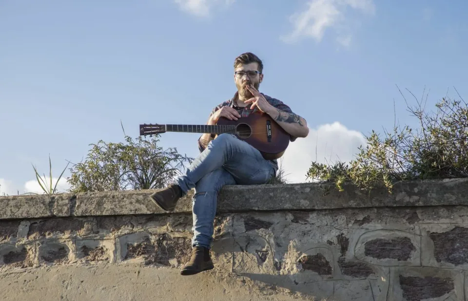 Matteo Leone seduto su un muretto di pietra con una chitarra acustica, cielo azzurro
